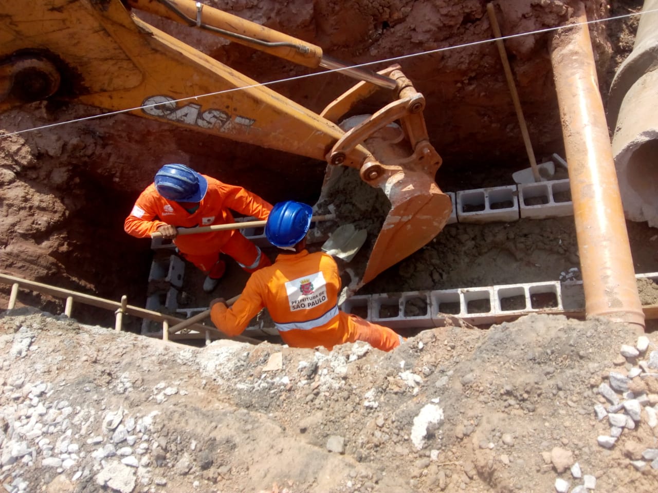 Dois funcionários dentro de um buraco retiram cimento de escavadeira para empilhar blocos de concreto.